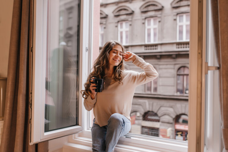 Refined caucasian girl in trendy shirt enjoying city views from window. Attractive curly woman chilling at home, sitting on sill with smile.